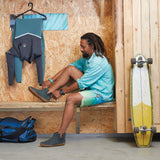 Man sitting on a bench with a surfboard next to him, wearing a green shirt and shorts with green ankle boots with honey rubber soles.