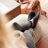 Person wearing black shoes and gray pants sitting on a textured stool with a brown leather bag in the background.