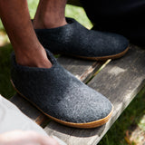 Person wearing dark gray slippers with honey rubber soles on a wooden surface outdoors.