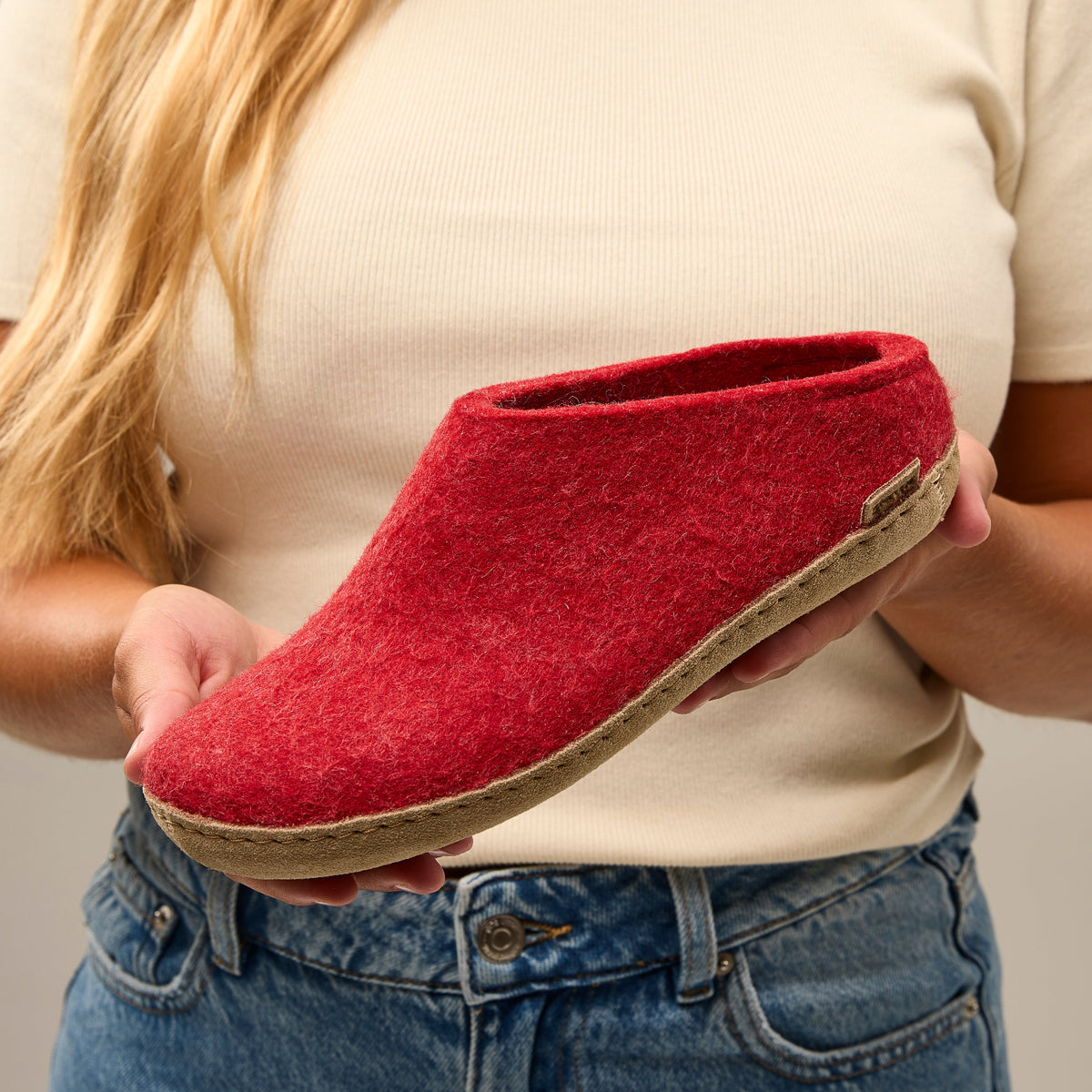 Red slipper held by a person wearing a beige shirt and blue jeans.
