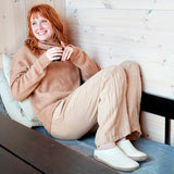 Woman sitting on a couch wearing a beige sweater and pants, holding a cup wearing white slippers