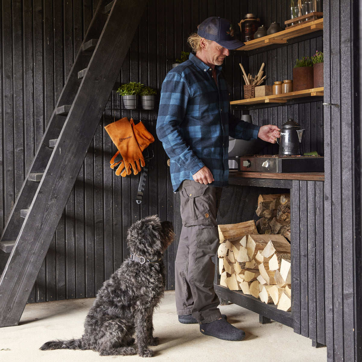 Man in a blue plaid shirt and gray pants standing next to a dog in a rustic cabin setting.
