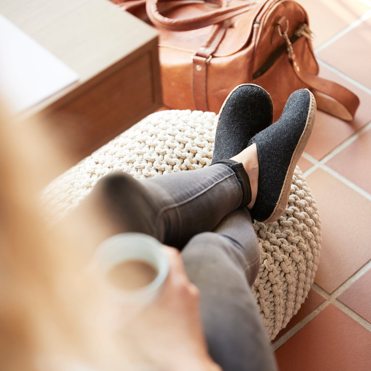 Person wearing black shoes and gray pants sitting on a textured stool with a brown leather bag in the background.