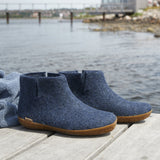 Pair of blue ankle boots with honey rubber  soles on a wooden dock by a body of water.
