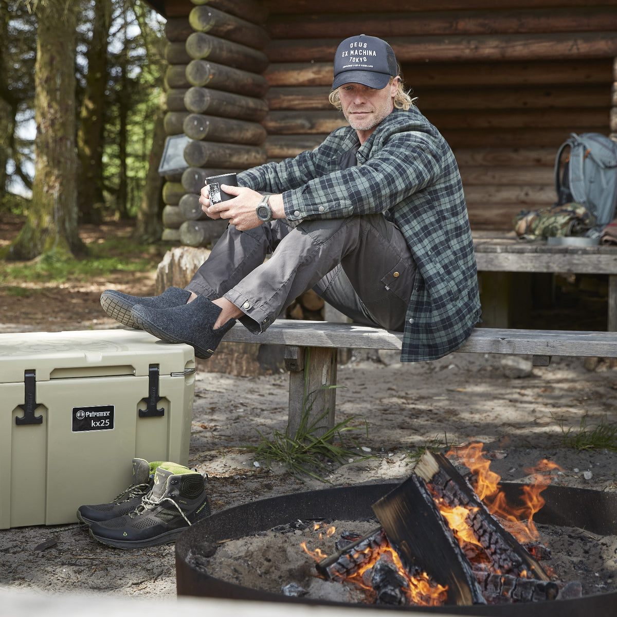 Man sitting by a campfire with a cooler and boots nearby, in front of a wooden cabin.