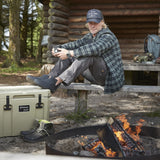 Man sitting by a campfire with a cooler and boots nearby, in front of a wooden cabin.