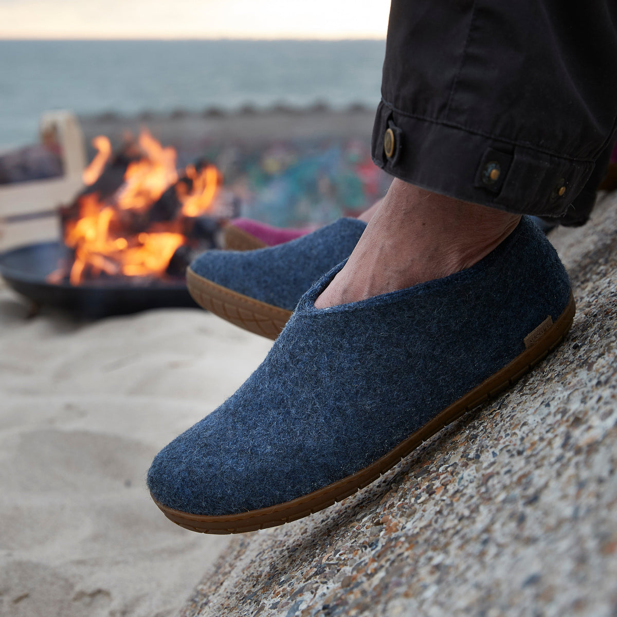 Person wearing blue slippers on a beach with a fire pit in the background