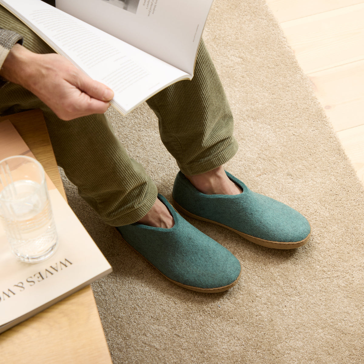 Person wearing North Sea slippers sitting on a couch with a glass of water and book.