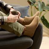 Person sitting on a couch wearing beige boots and holding a mug, with a plant in the background.