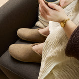 Person wearing sand shoes and holding a cup, wrapped in a beige blanket on a couch.