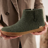 Green ankle boot with  honey rubber sole held by a person wearing a beige coat.