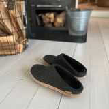 Forest green slippers on a wooden floor with a fireplace in the background