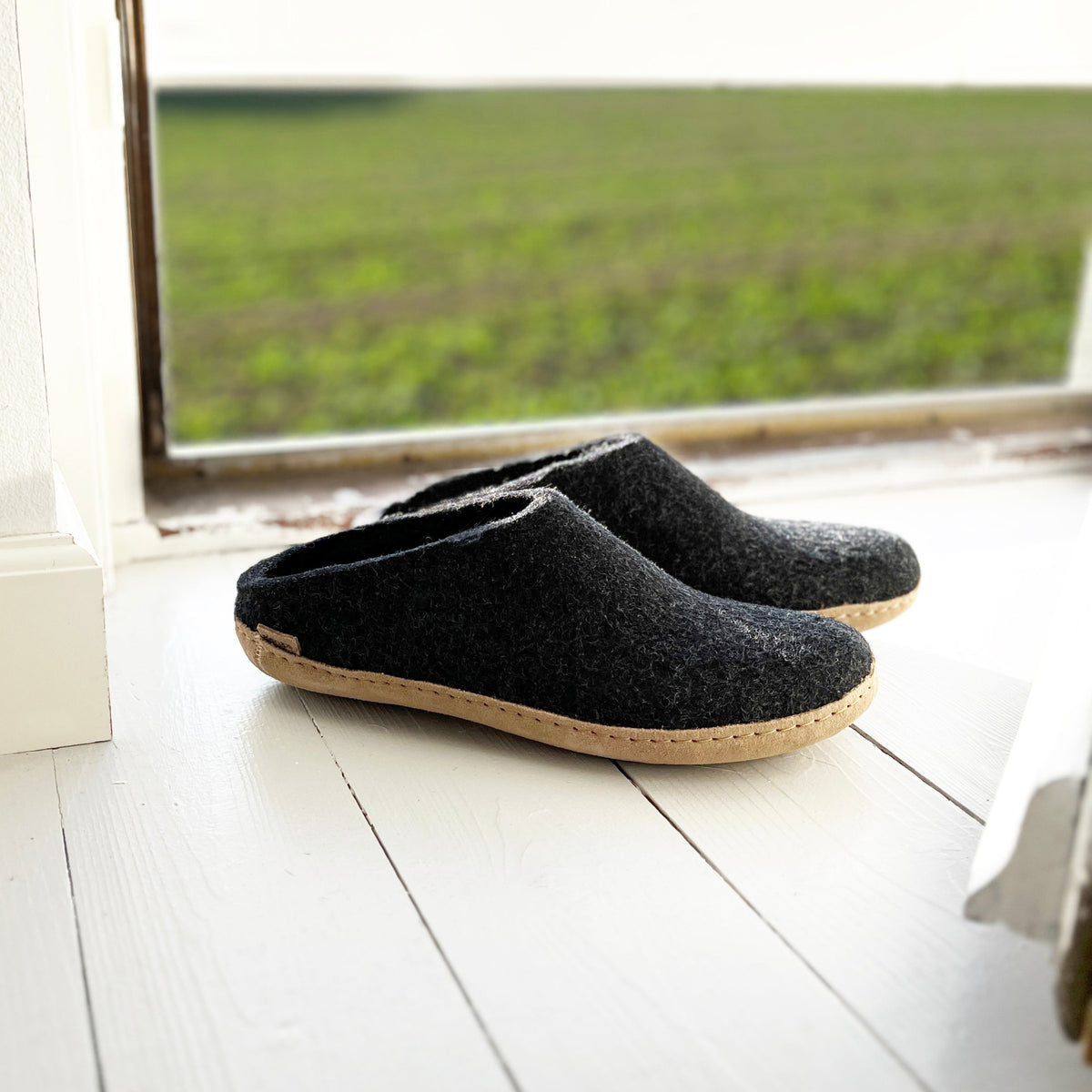 Pair of dark gray slippers on a light wooden floor with a window in the background showing greenery.